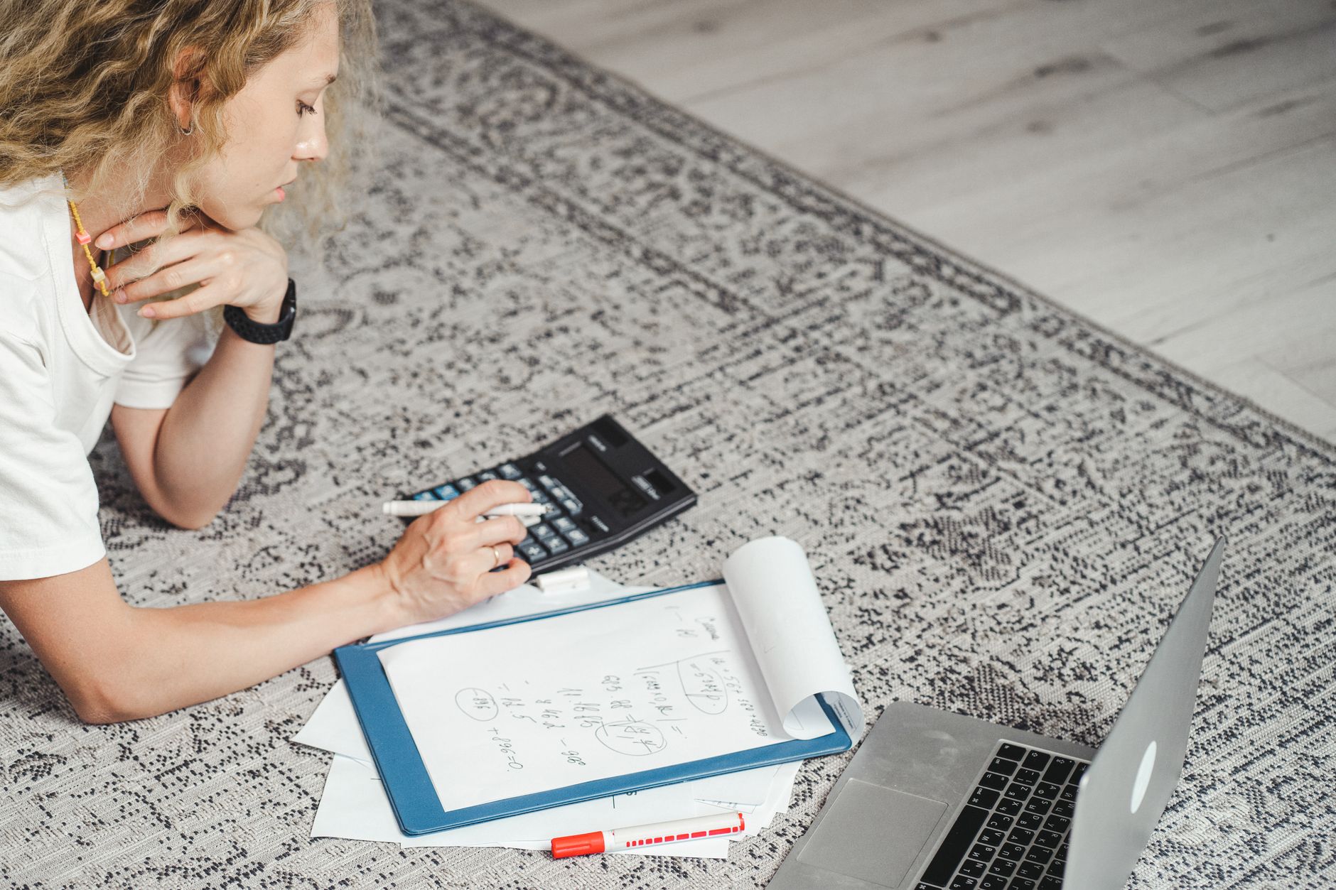 Une femme calcule ses finances à la maison à l'aide d'un ordinateur portable, d'une calculatrice et de documents sur un tapis.