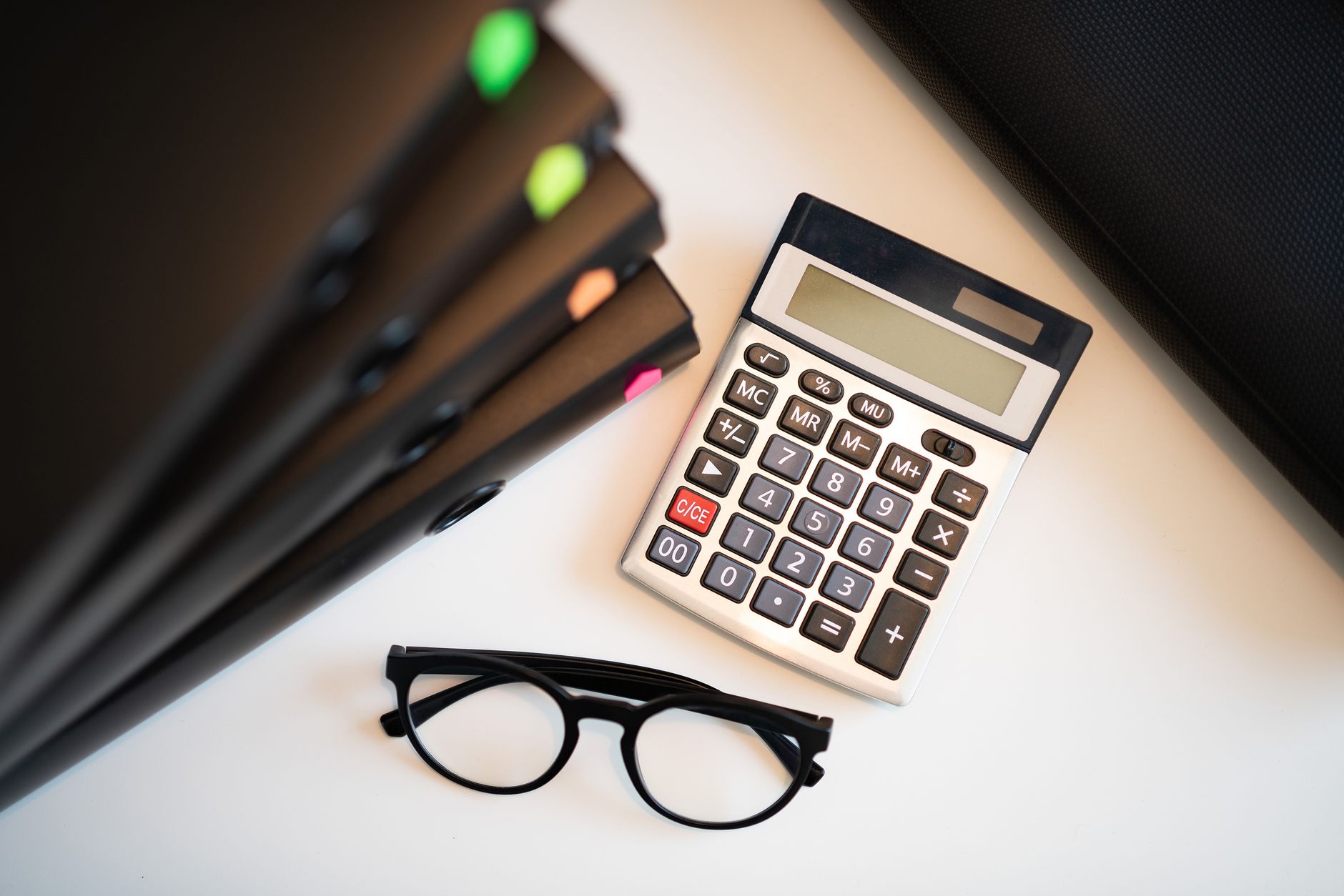 Calculatrice, lunettes et classeurs posés sur un bureau blanc