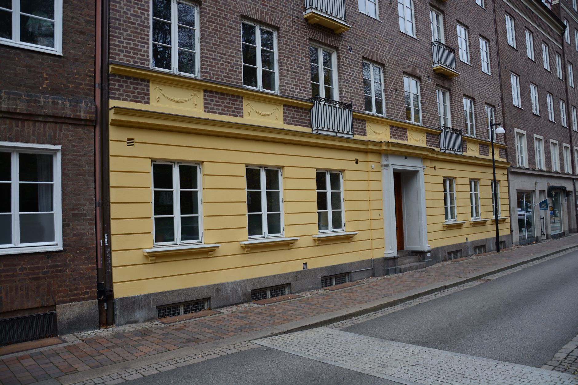 Façade d’un immeuble en brique au rez-de-chaussée jaune, avec fenêtres blanches, balcons en fer forgé et trottoir pavé le long d’une rue.