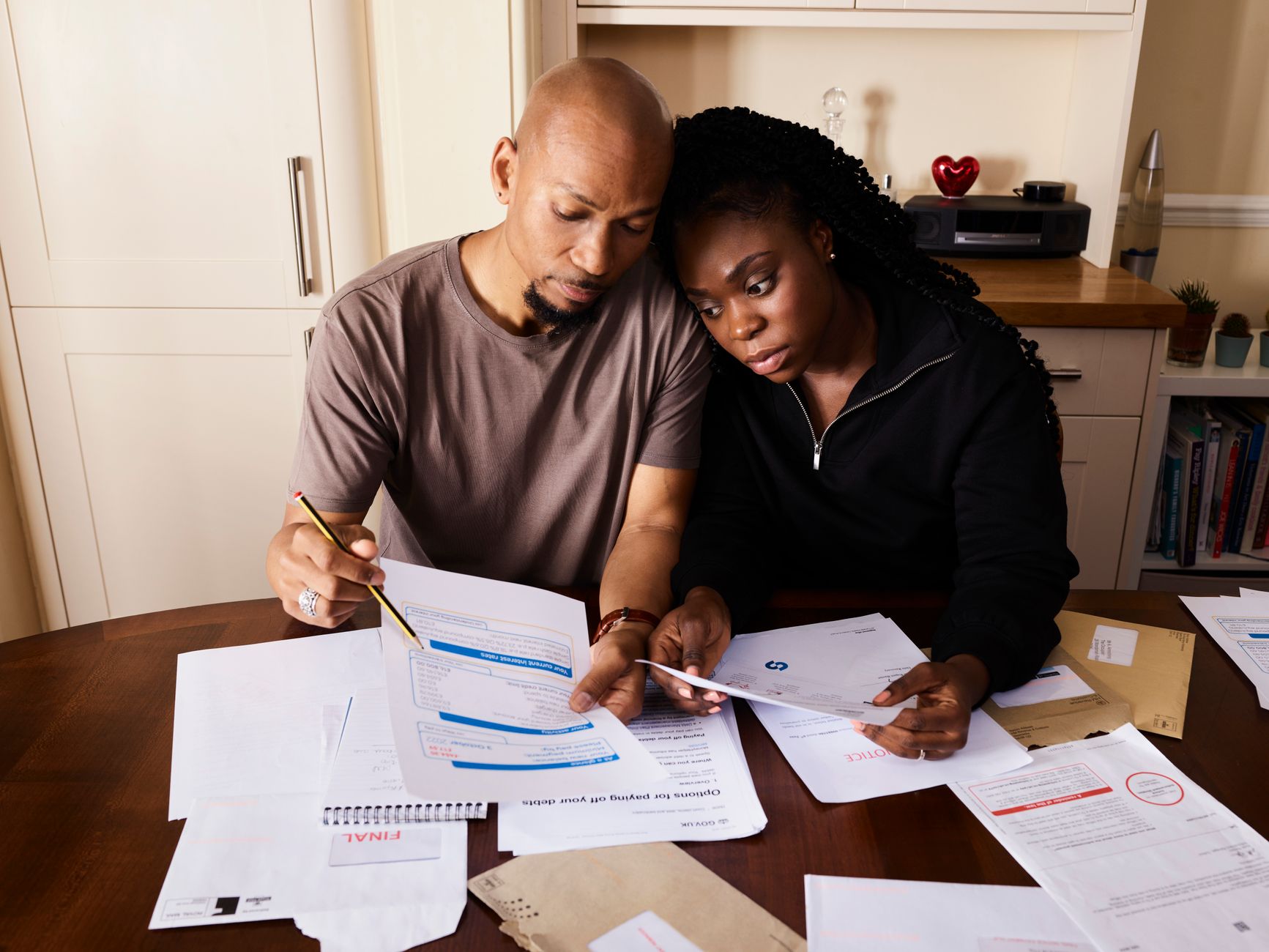 Un couple examine des documents papier étalés sur une table en bois dans une cuisine, l’air concentré.